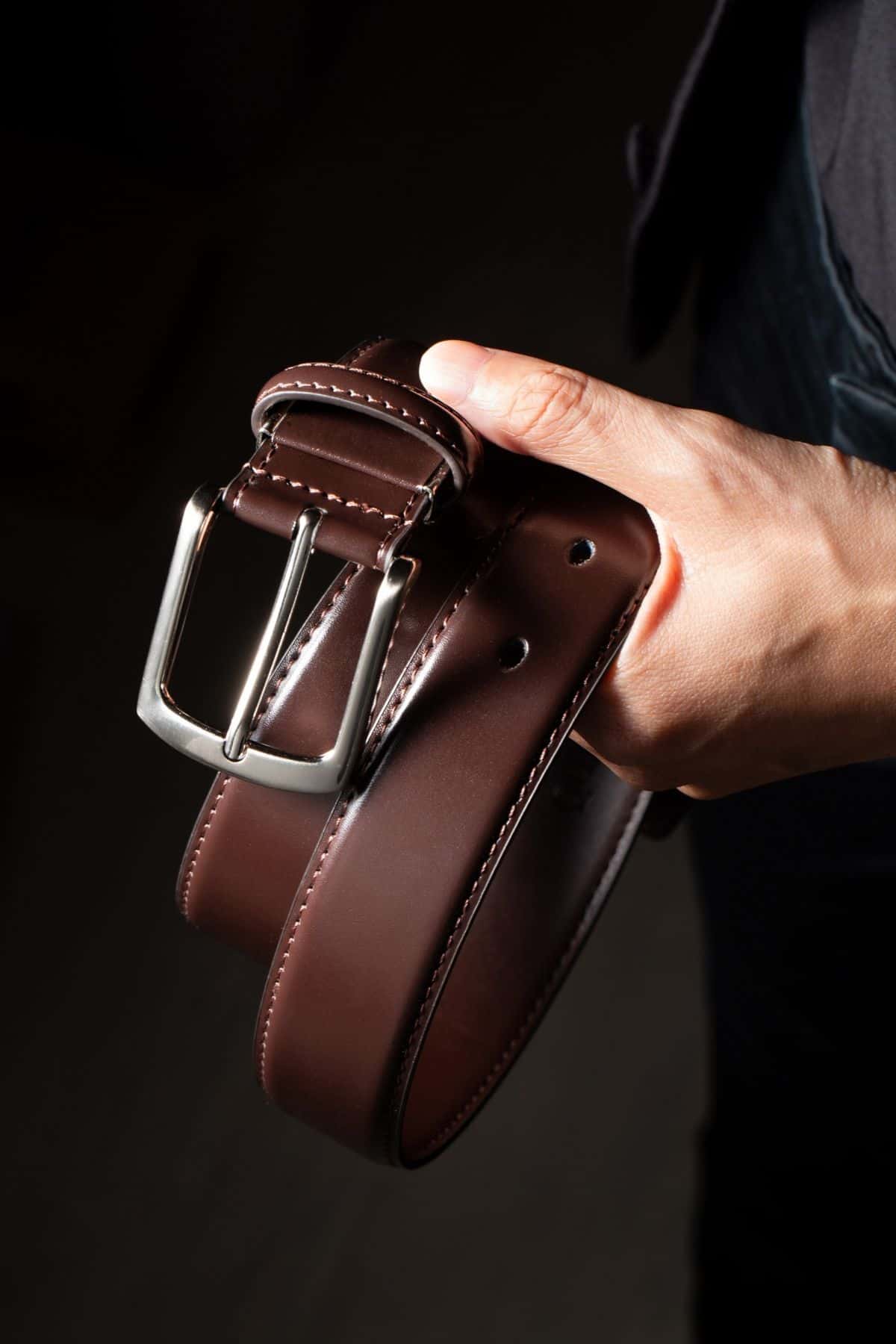 A hand holding a brown leather belt with a brushed pin buckle