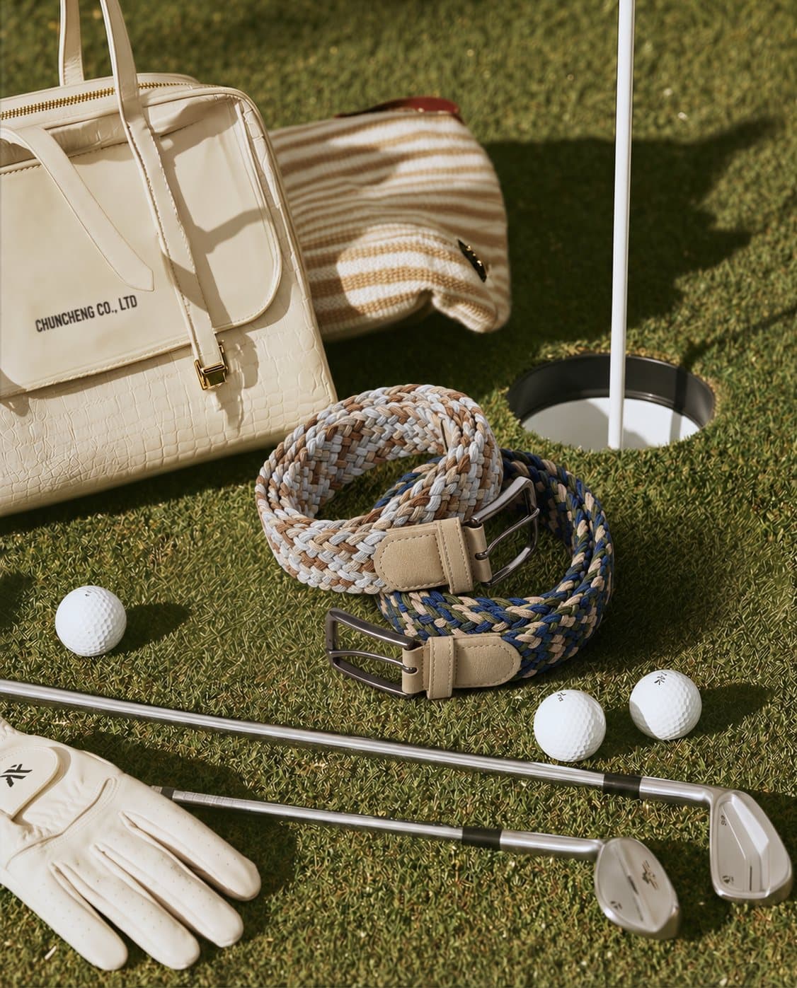 Two braided stretch belts on a golf green beside clubs, balls and a white leather bag
