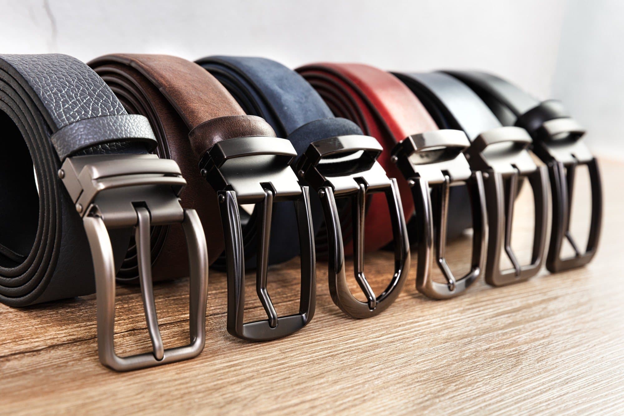 Six leather pin-buckle belts in a row — pebble grain, brown, navy suede, red, grey and black